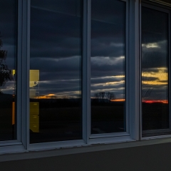 A long window divided into narrow panes reflects clouds and a sunset sky with a red streak along the horizon, with silhouetted trees. We see what might be a filing cabinet and a tiny print of a hand, perhaps on the wall above the filing cabinet.